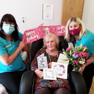Lady resident with cards and flowers on her birthday at Hengist Field Care Home, with two staff members holding a birthday banner